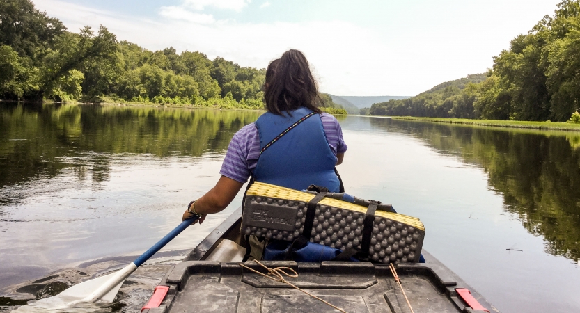 From the back of a canoe, a person in front paddles a canoe on calm water framed by green trees. 